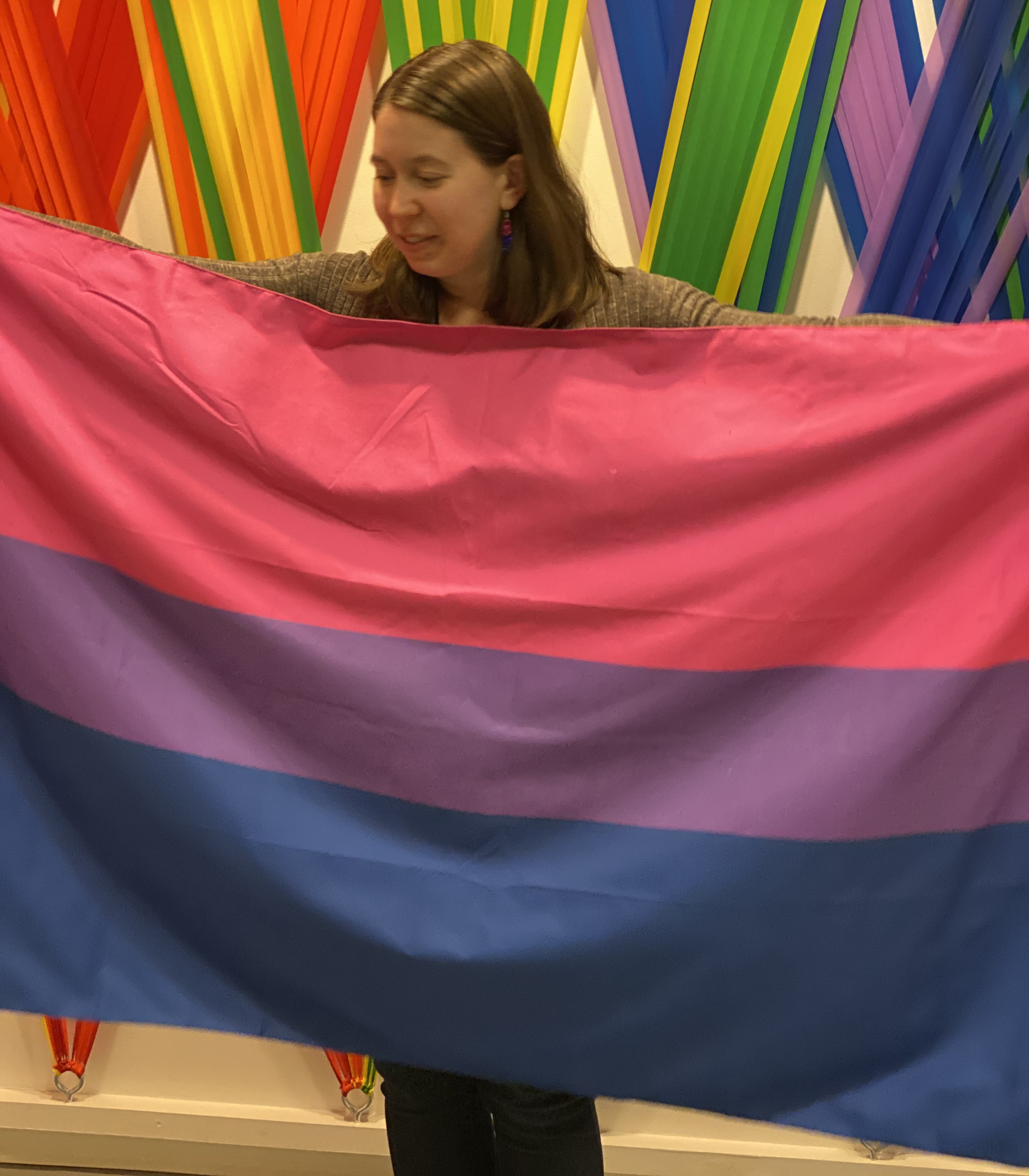 M holds a bi flag (pink stripe, purple stripe, blue stripe) in front of themself and looks down at the flag. Rainbow decorations are behind them.