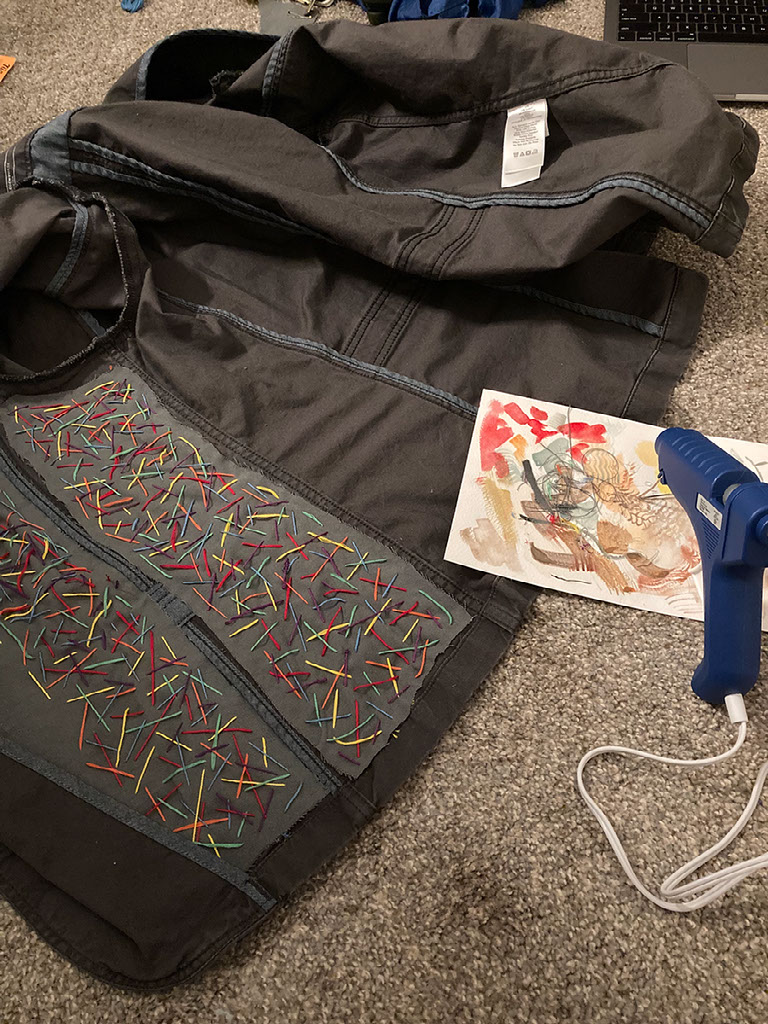the inside of the jacket, now with two embroidered panels glued in. a blue hot glue gun is also pictured. Everything is on the floor