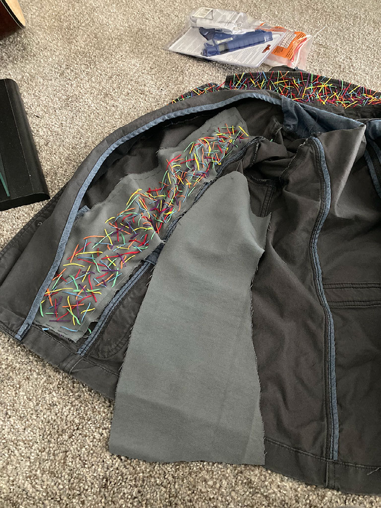 A jacket on the floor, opened to the inside. There is one embroidered panel messily stitched in. The lining puckers. A blue hot glue gun is on the top of the image
