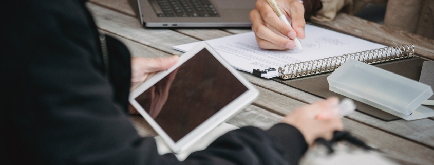 A pari of hands holding a tablet across the table from another pair of hands writing in a notebook