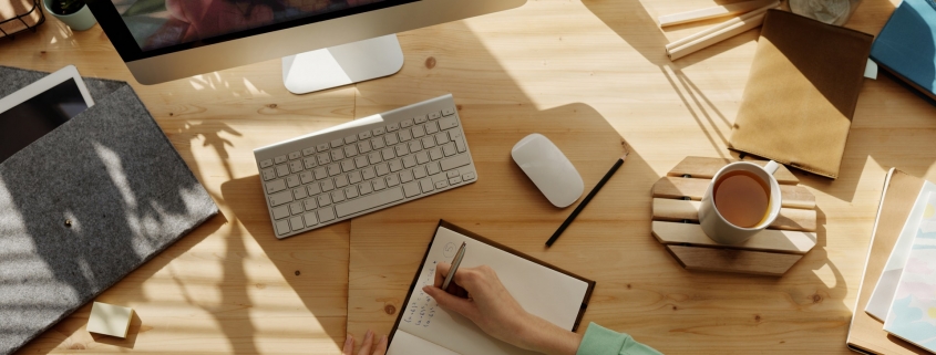 Desk with a person taking notes in a book while a computer shows a woman's face on video chat